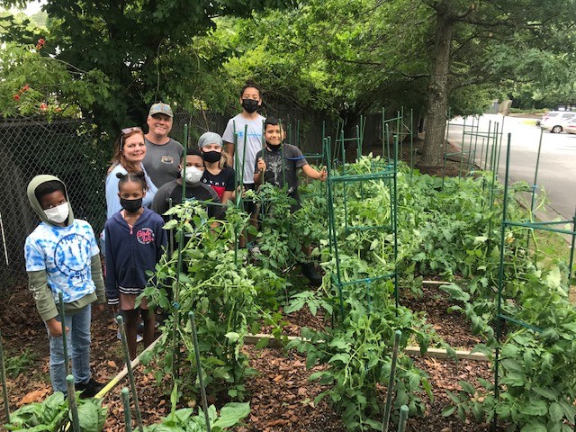 Group of children and adults stand together in a garden surrounded by tall tomato plants and green supports.