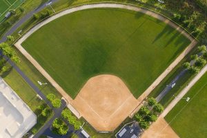 Aerial view of an empty baseball field with green grass, a dirt infield, and surrounding paths and trees. Benches and bleachers are visible along the sides.