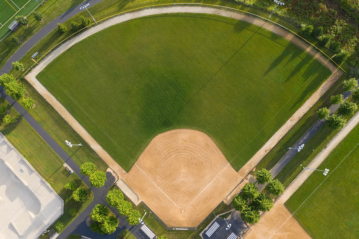Aerial view of an empty baseball field with green grass, a dirt infield, and surrounding paths and trees. Benches and bleachers are visible along the sides.
