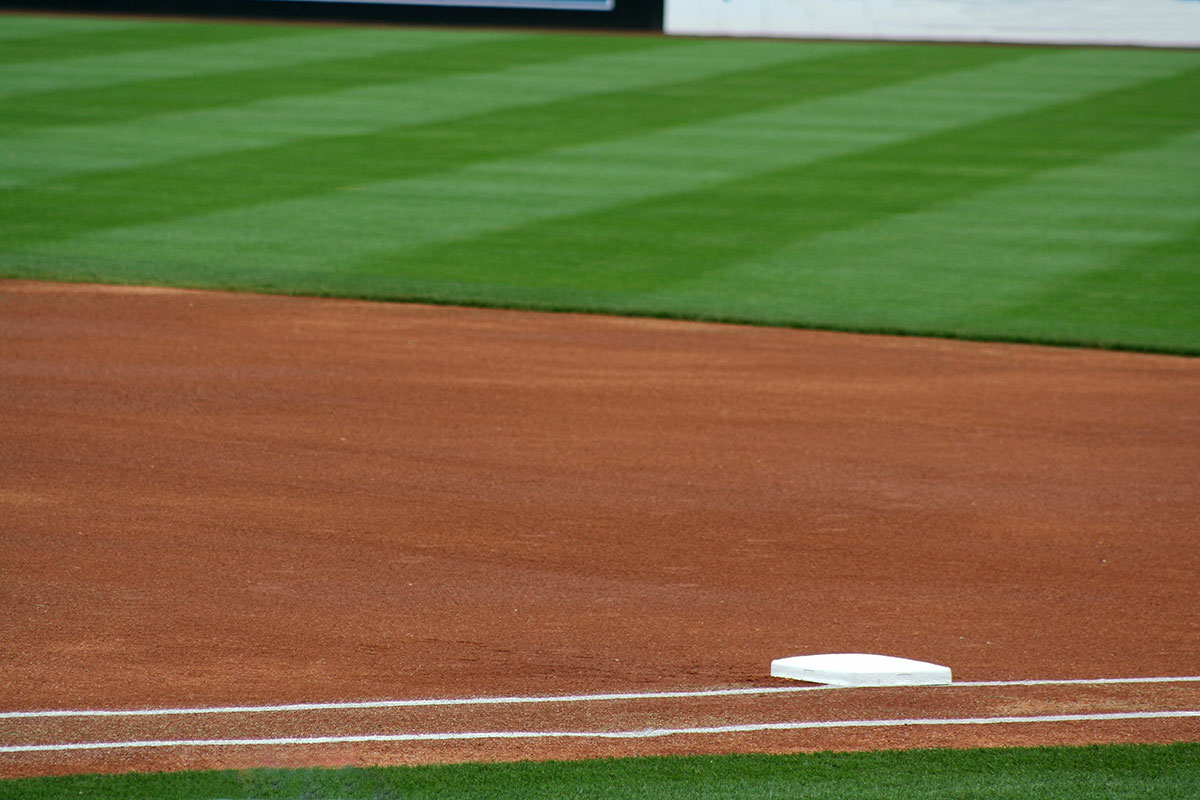 A close-up view of a baseball field showing the dirt infield, green grass outfield, and a white base near the bottom right corner.