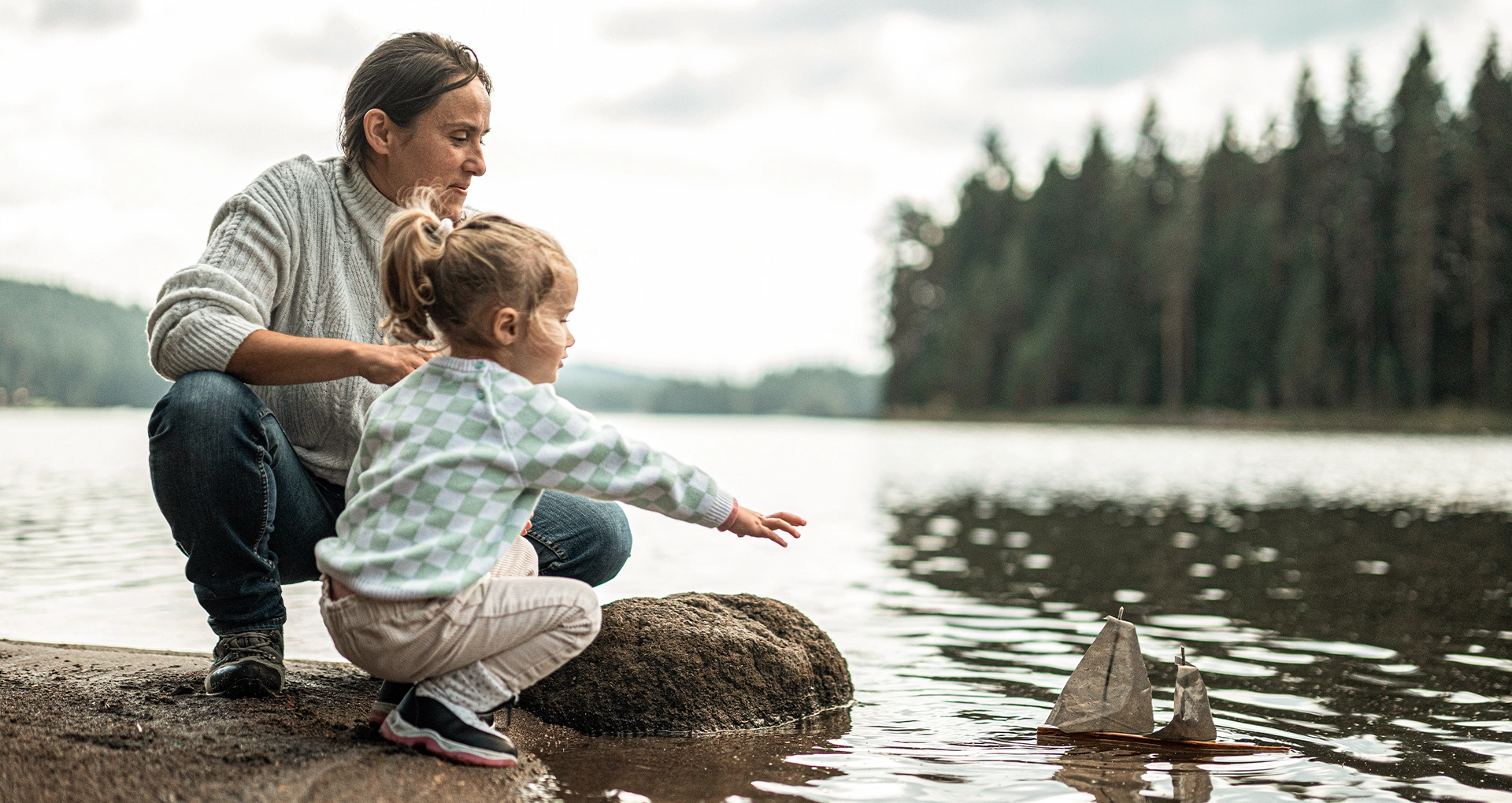 An adult and a young child kneel by a lake, watching a small toy sailboat float on the water. Trees line the shore in the background.