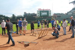 A group of people gathers on a baseball field near wooden equipment and tools, with a scoreboard and empty stadium seats visible in the background.