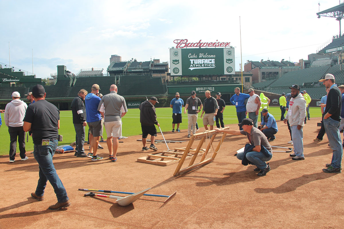 A group of people gathers on a baseball field near wooden equipment and tools, with a scoreboard and empty stadium seats visible in the background.