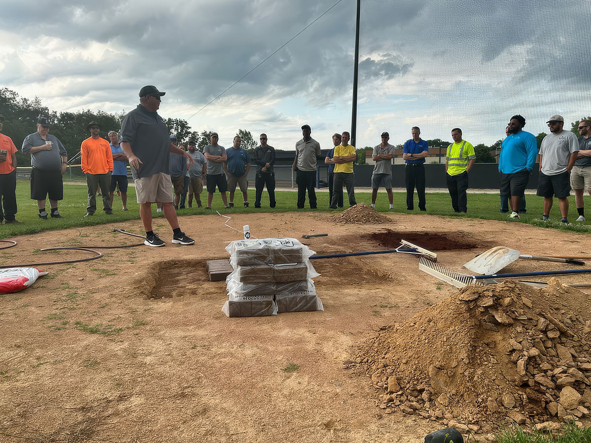 A man speaks to a group of people standing in a semi-circle on a baseball field. Tools, dirt piles, and bags of material are on the ground near him. The sky is cloudy.