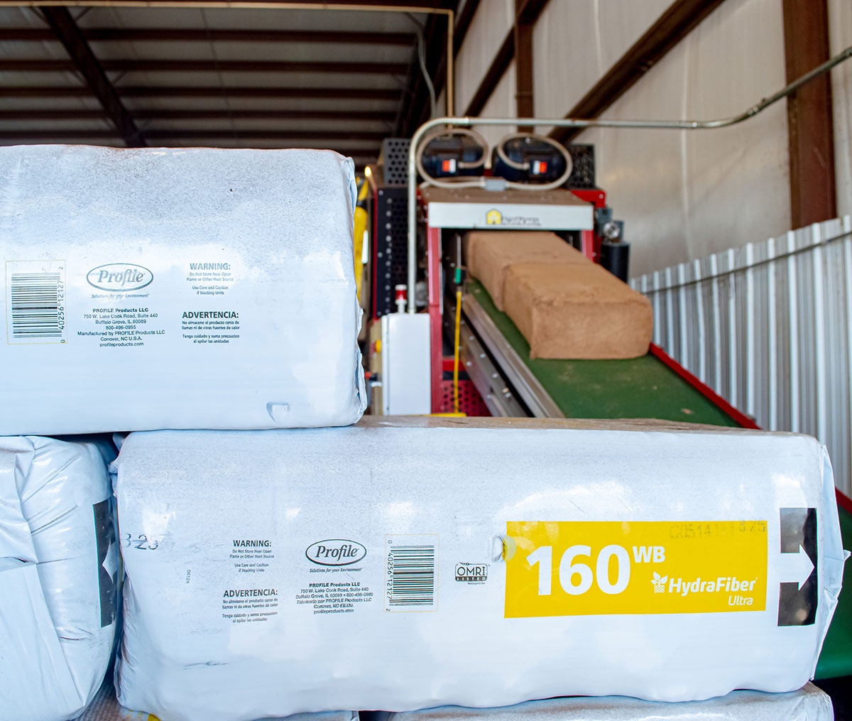 Stacks of compressed HydraFiber Ultra 160WB bales sit near a green conveyor belt inside an industrial warehouse with metal walls and ceiling.