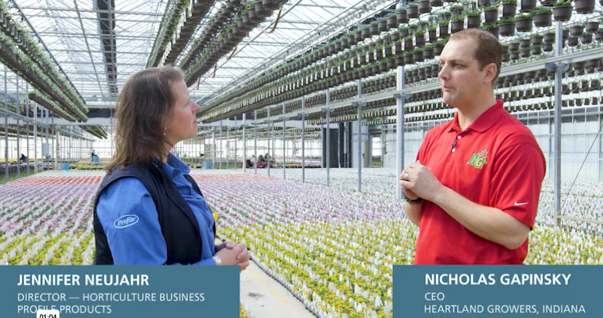 Two people converse in a large greenhouse filled with rows of potted plants. Both are standing; one wears a blue shirt and vest, and the other wears a red polo shirt. Text boxes display their names and titles.