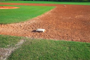 A close-up view of a baseball infield showing a base on the dirt, surrounded by green grass under bright sunlight. The field appears empty and well-maintained.