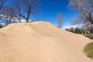 A large pile of light-colored sand sits in a park with bare trees and blue sky in the background.