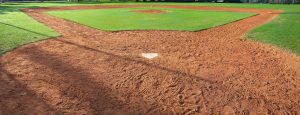 A baseball field with home plate in the foreground, reddish dirt base paths, and green grass covering the pitchers mound and outfield under bright sunlight.