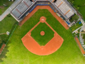 Aerial view of an empty baseball field, showing the diamond, bases, pitchers mound, grassy outfield, and empty bleachers on both sides.
