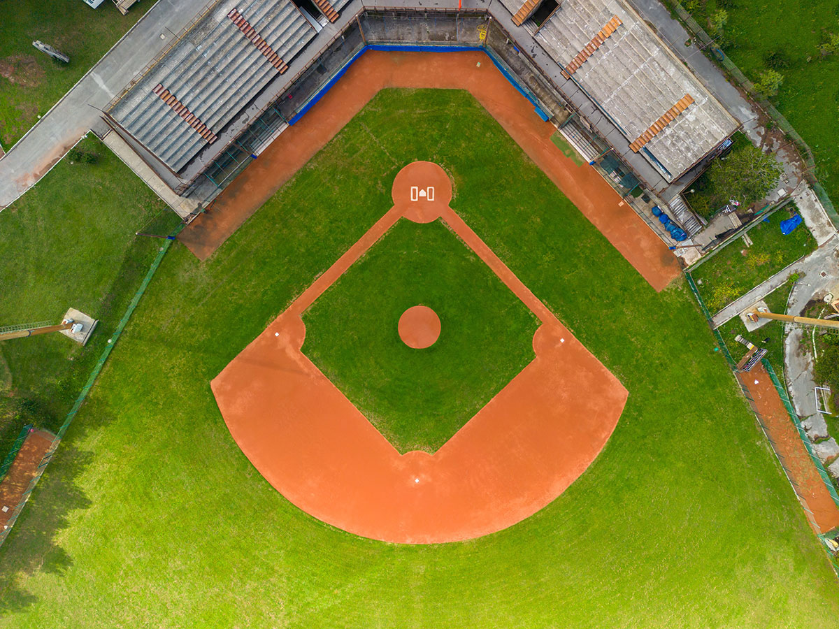 Aerial view of an empty baseball field, showing the diamond, bases, pitchers mound, grassy outfield, and empty bleachers on both sides.