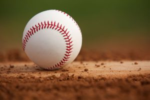 A white baseball with red stitching rests on a dirt pitchers mound, with a green outfield blurred in the background.