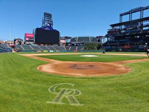 View from behind home plate at an empty baseball stadium with a large video scoreboard and the CR logo on the grass, under a clear blue sky.