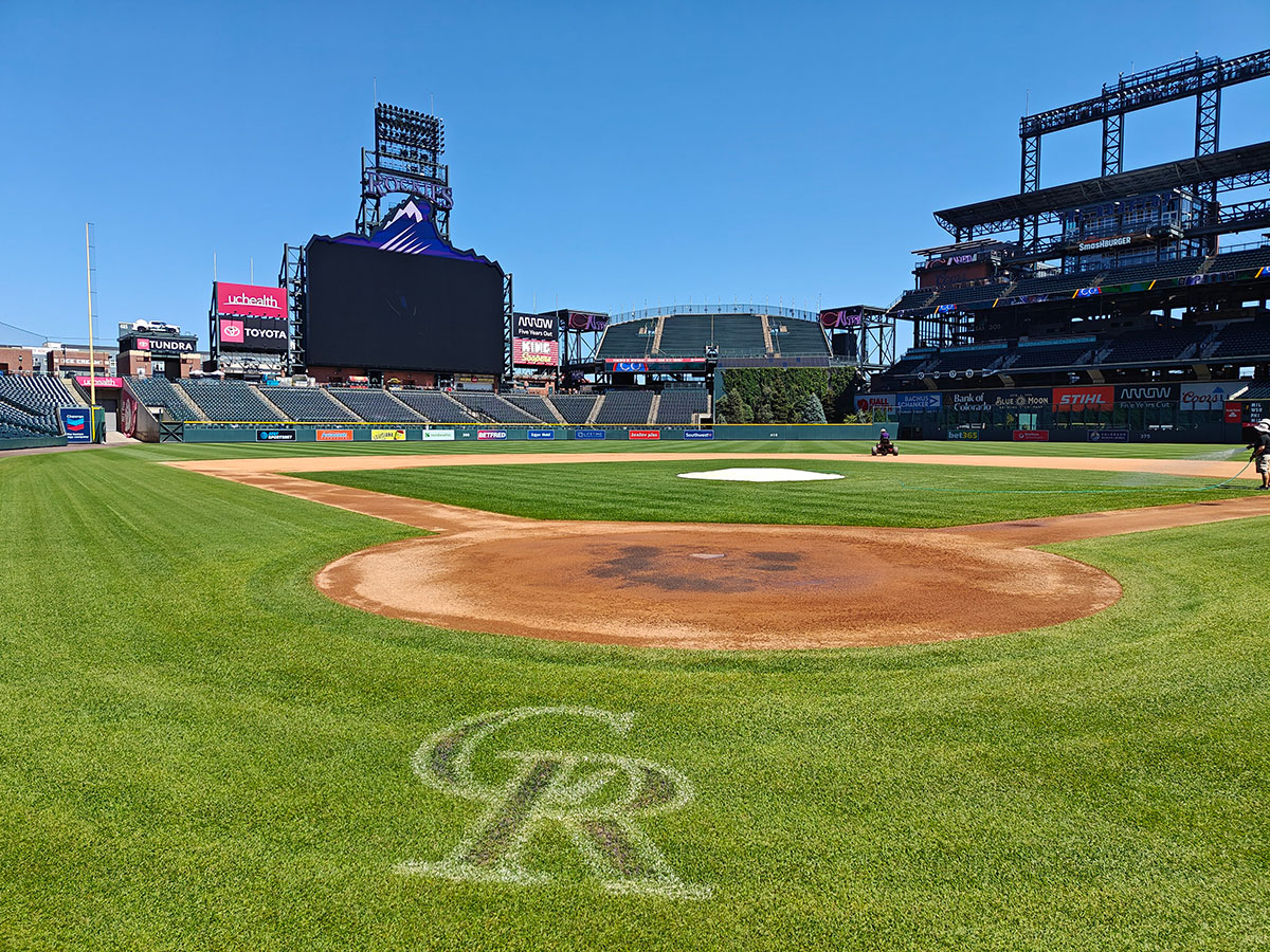 View from behind home plate at an empty baseball stadium with a large video scoreboard and the CR logo on the grass, under a clear blue sky.