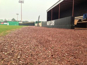 A low-angle view of a baseball field shows the gravel path next to the dugout, with green grass and fences visible in the distance under a cloudy sky.
