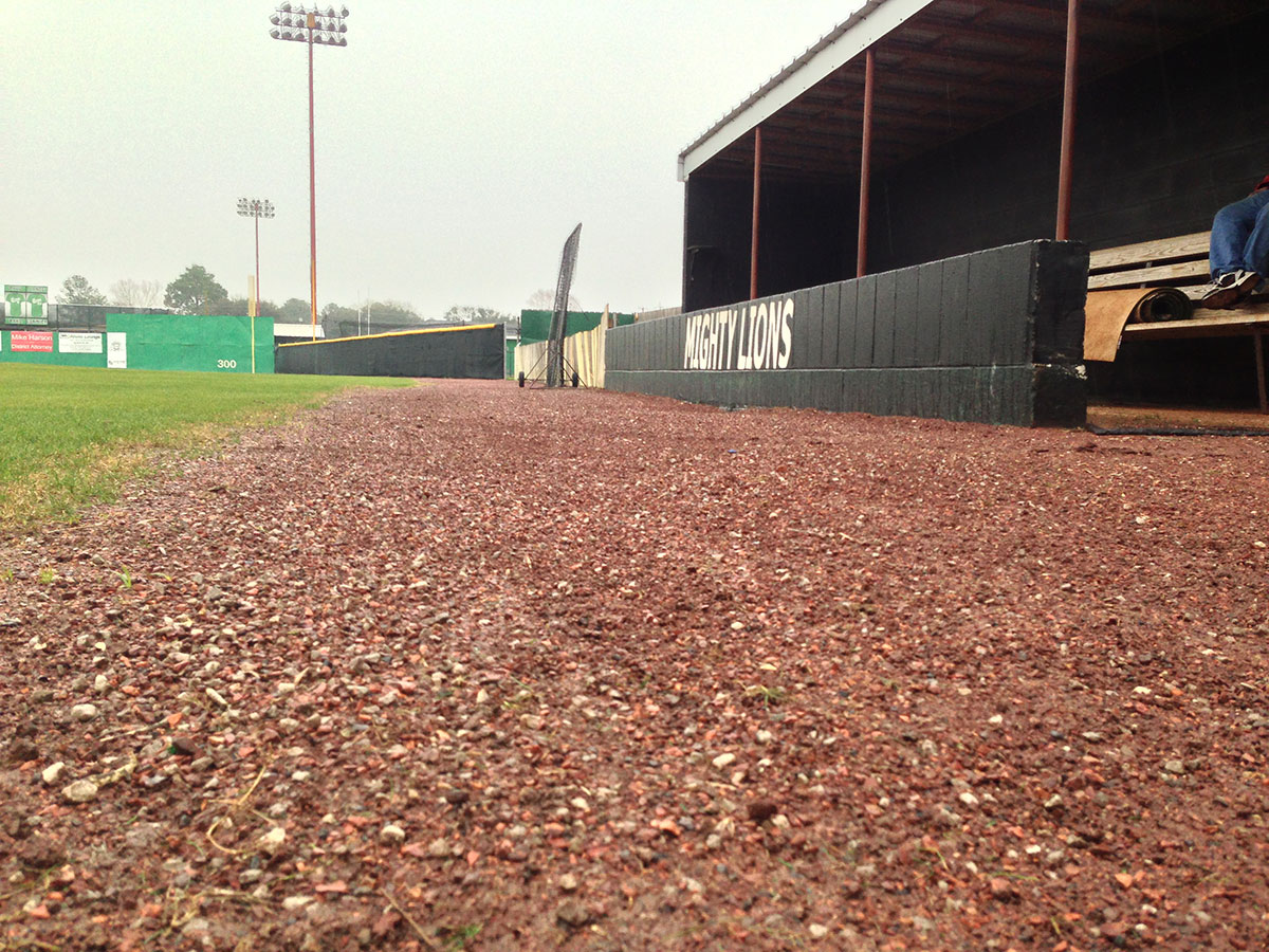 A low-angle view of a baseball field shows the gravel path next to the dugout, with green grass and fences visible in the distance under a cloudy sky.