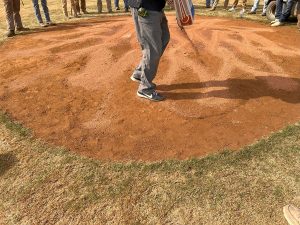 A person stands on a baseball pitchers mound made of reddish dirt, surrounded by others whose lower legs and shoes are visible, suggesting a group gathering or sports event.