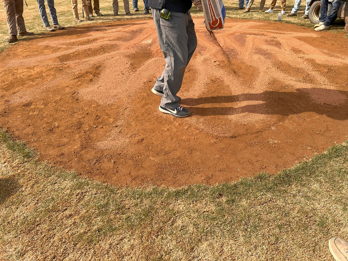 A person stands on a baseball pitchers mound made of reddish dirt, surrounded by others whose lower legs and shoes are visible, suggesting a group gathering or sports event.