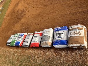 Seven large bags of various grass seed blends are lined up on a grassy and dirt field, each with different colored packaging and labels, ready for planting or landscaping use.