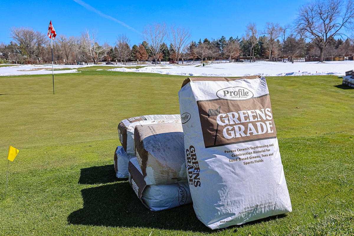Large bags labeled Greens Grade sit on the green of a golf course. Snow covers parts of the surrounding landscape, and trees and a flagstick are visible in the background.