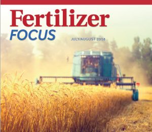 A blurred blue harvester works in a golden wheat field under a hazy sky. The magazine cover reads Fertilizer Focus and July/August 2018..