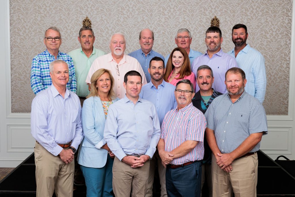 A group of fifteen adults, mostly men with one woman, pose together indoors in two rows, all smiling at the camera. They are dressed in business-casual attire against a patterned wall.