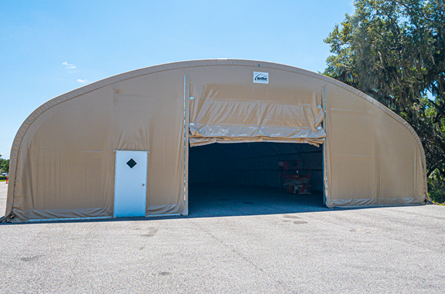 A large tan fabric-covered storage building with a wide open entrance and a white side door sits on a paved lot under a clear blue sky.