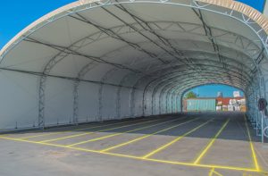 A large open-sided fabric storage tent with a metal frame, covering a paved area marked with yellow lines. Shipping containers and stacked materials are visible in the background.