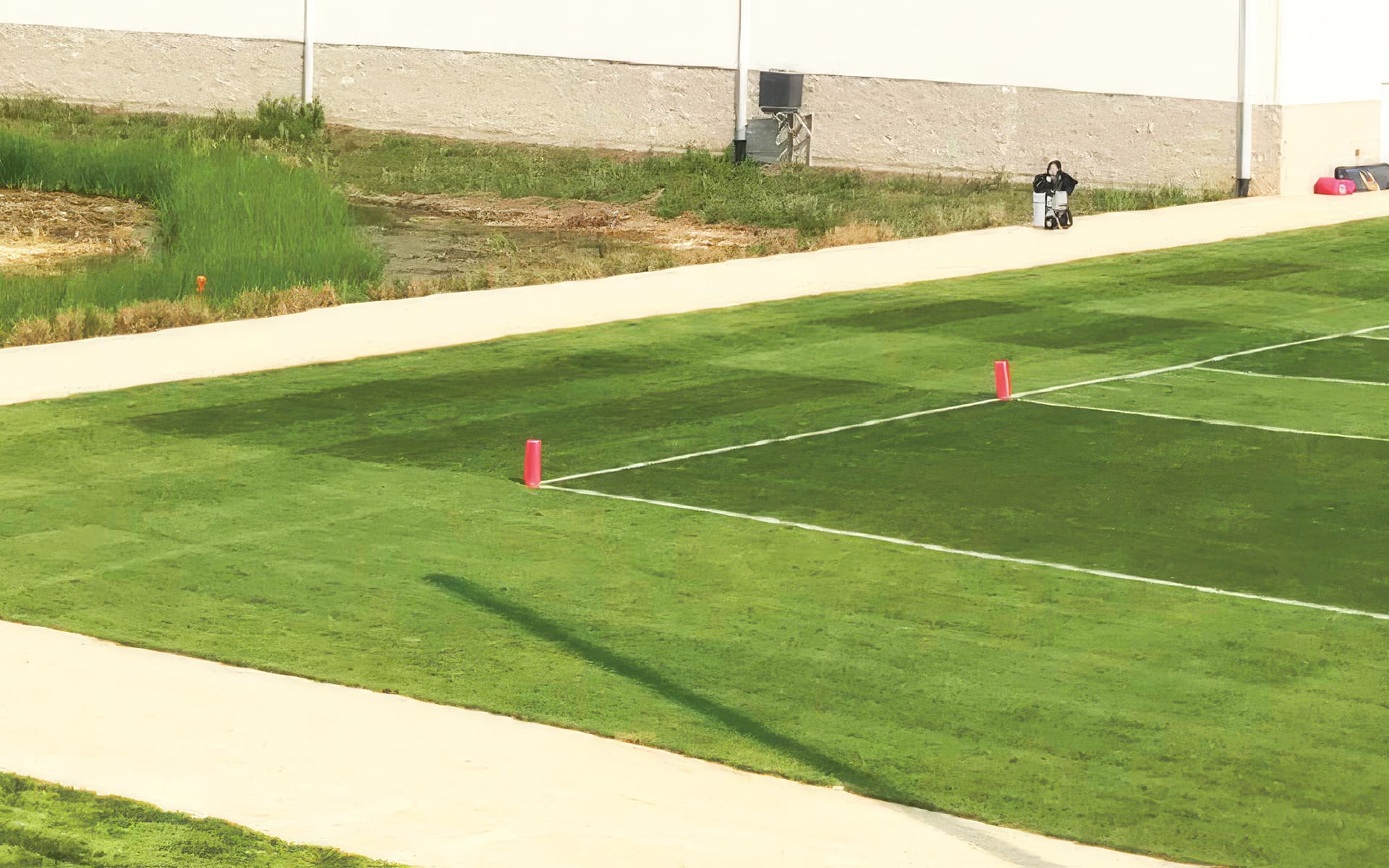 A person rides a lawn mower along a sidewalk next to a green sports field marked with white lines and pink markers.