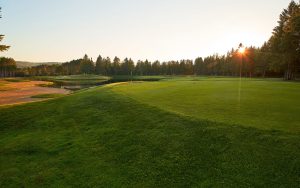 A golf course at sunset, with green fairways, trees in the background, a sand trap to the left, and sunlight streaming through the trees on the right.
