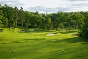 A lush green golf course with sand bunkers, surrounded by trees under a partly cloudy sky.