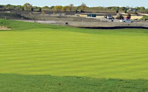 A bright green golf course with mowed stripes is in the foreground. In the background, there is construction equipment, vehicles, and bare soil, showing ongoing development behind the grass. Trees line the horizon.