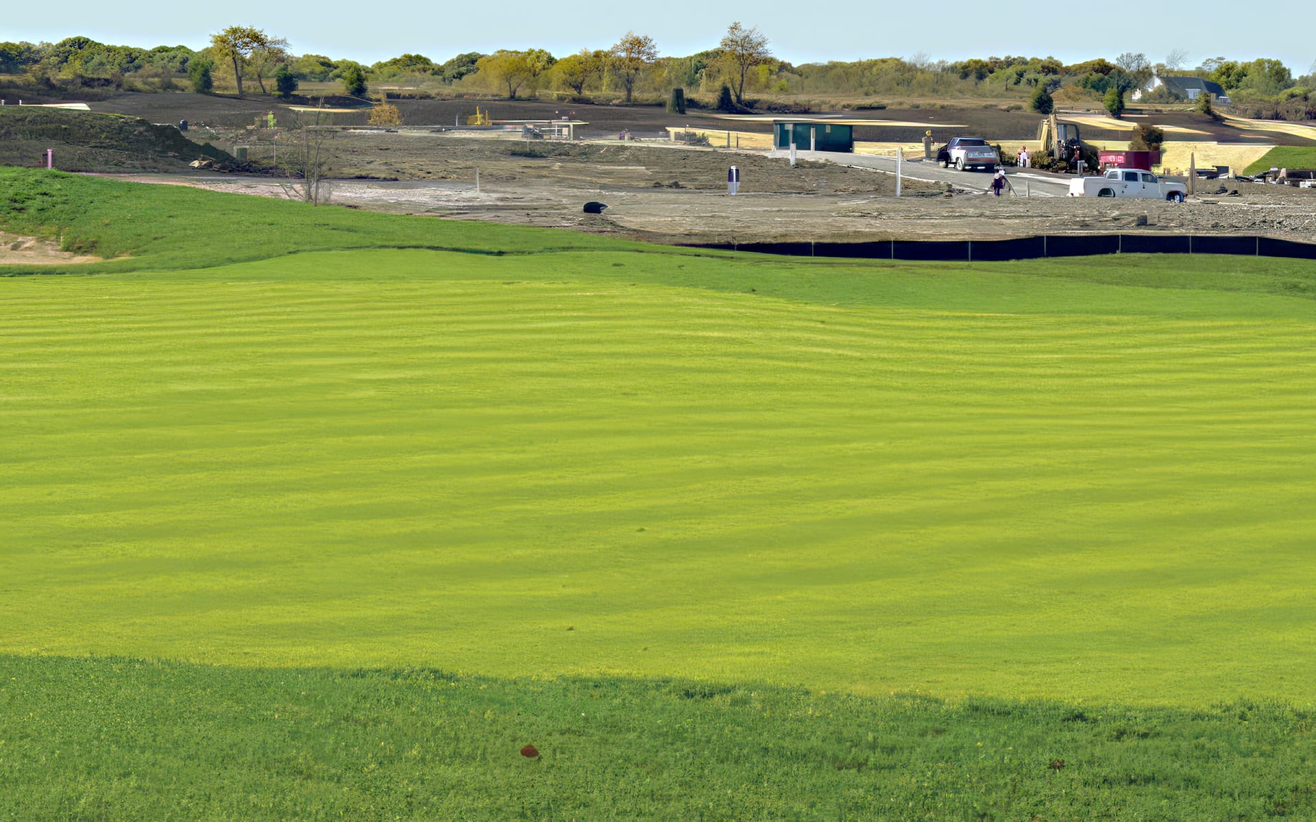A bright green golf course with mowed stripes is in the foreground. In the background, there is construction equipment, vehicles, and bare soil, showing ongoing development behind the grass. Trees line the horizon.