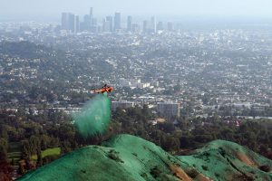 A helicopter sprays green material over hills, likely for erosion control, with a sprawling city and tall buildings in the background.