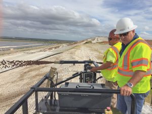 Two men in safety gear operate equipment on a sandy construction site, overseeing a machine dispersing material on a slope with cloudy skies and distant water visible in the background.