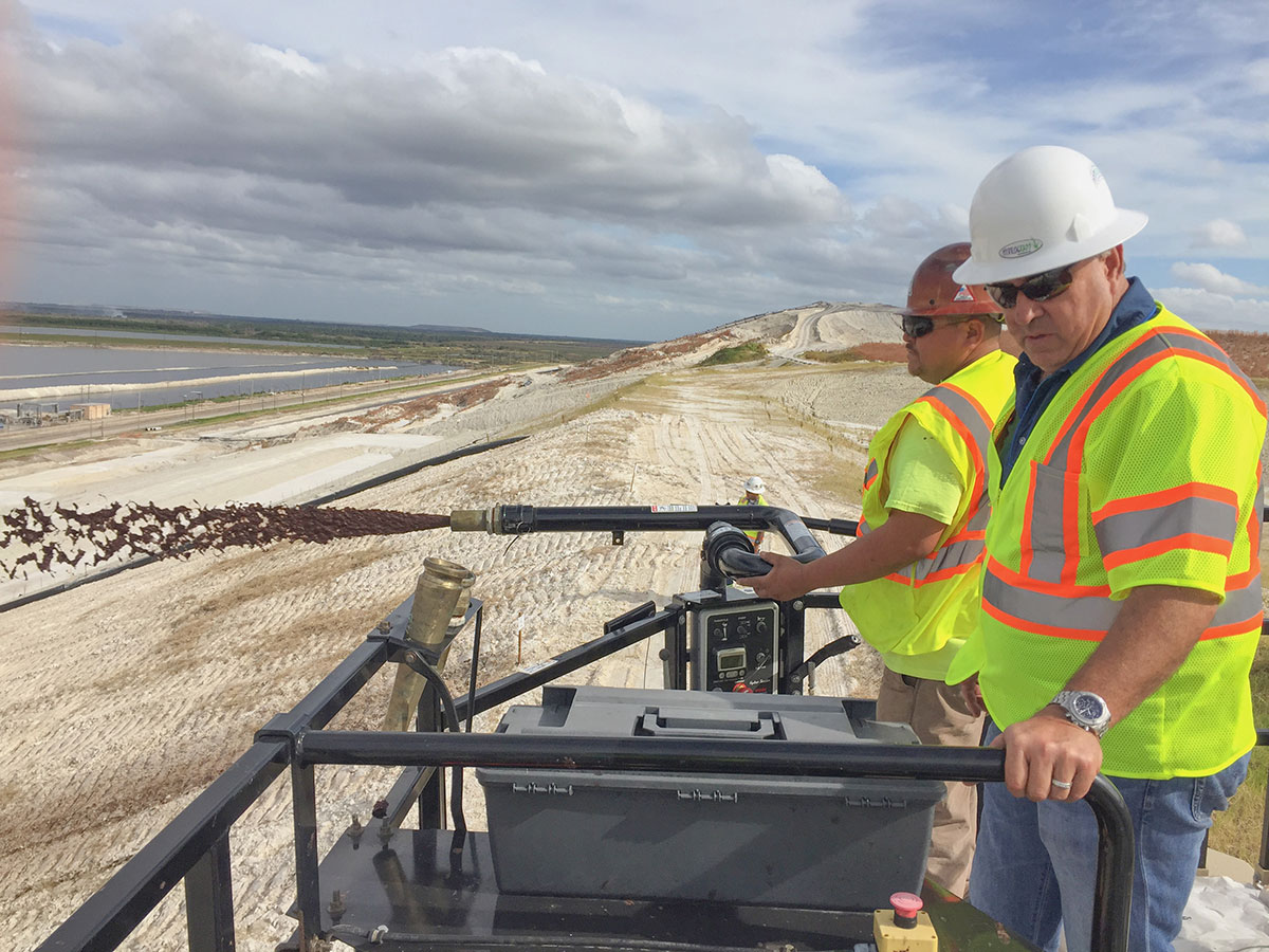 Two men in safety gear operate equipment on a sandy construction site, overseeing a machine dispersing material on a slope with cloudy skies and distant water visible in the background.
