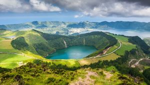 A lush green crater surrounds a blue lake, with winding roads and distant hills under a cloudy sky, overlooking a coastal town and the ocean in the background.