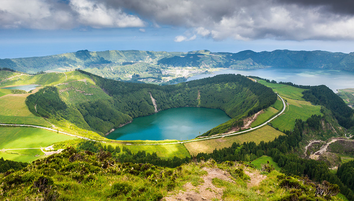 A lush green crater surrounds a blue lake, with winding roads and distant hills under a cloudy sky, overlooking a coastal town and the ocean in the background.