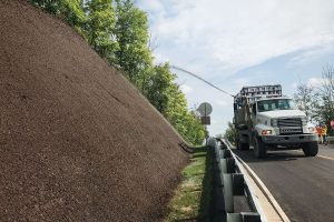 A truck sprays mulch onto a steep embankment beside a road, with trees in the background and a partly cloudy sky above.
