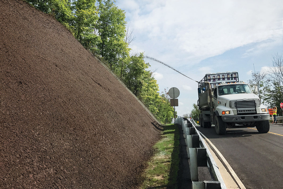 A truck sprays mulch onto a steep embankment beside a road, with trees in the background and a partly cloudy sky above.