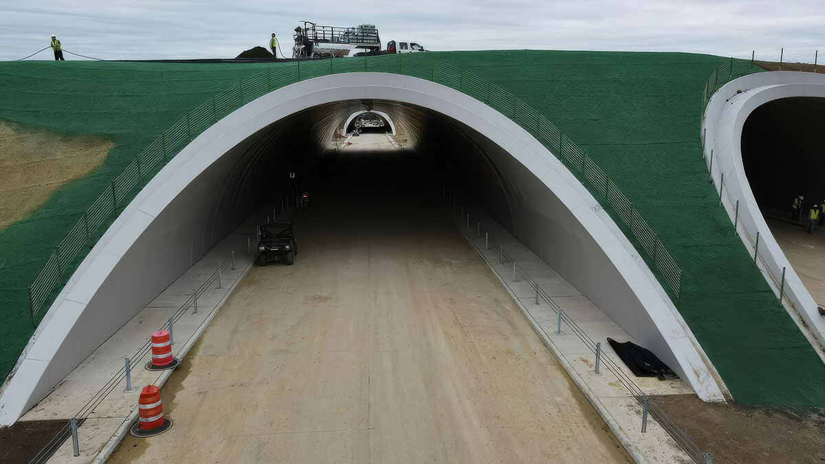 A wide road tunnel with arched entrances, covered by green artificial turf and lined with safety railings. Construction vehicles and workers are visible near and atop the tunnel. Orange cones block part of the entrance.