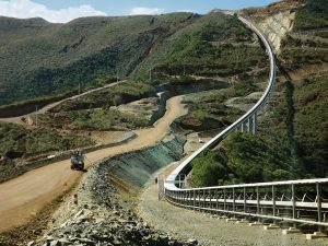 A winding dirt road and an elevated conveyor belt run parallel through a hilly, green landscape with construction vehicles visible along the route.