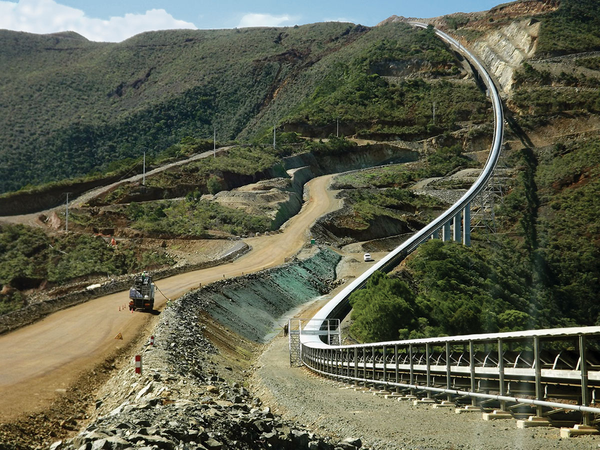 A winding dirt road and an elevated conveyor belt run parallel through a hilly, green landscape with construction vehicles visible along the route.