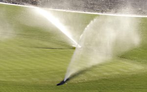 Two sprinklers spray water over a green, manicured lawn with a visible dirt border in the background. Sunlight highlights the water mist on the grass.