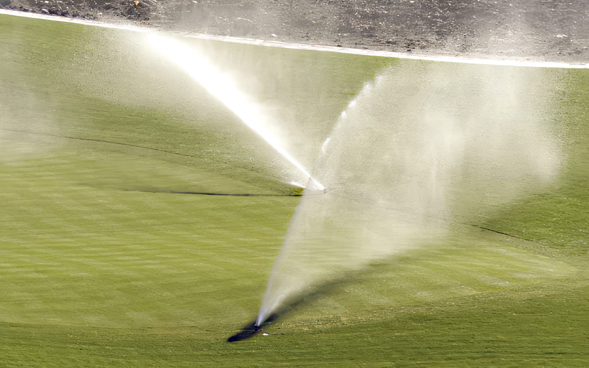 Two sprinklers spray water over a green, manicured lawn with a visible dirt border in the background. Sunlight highlights the water mist on the grass.
