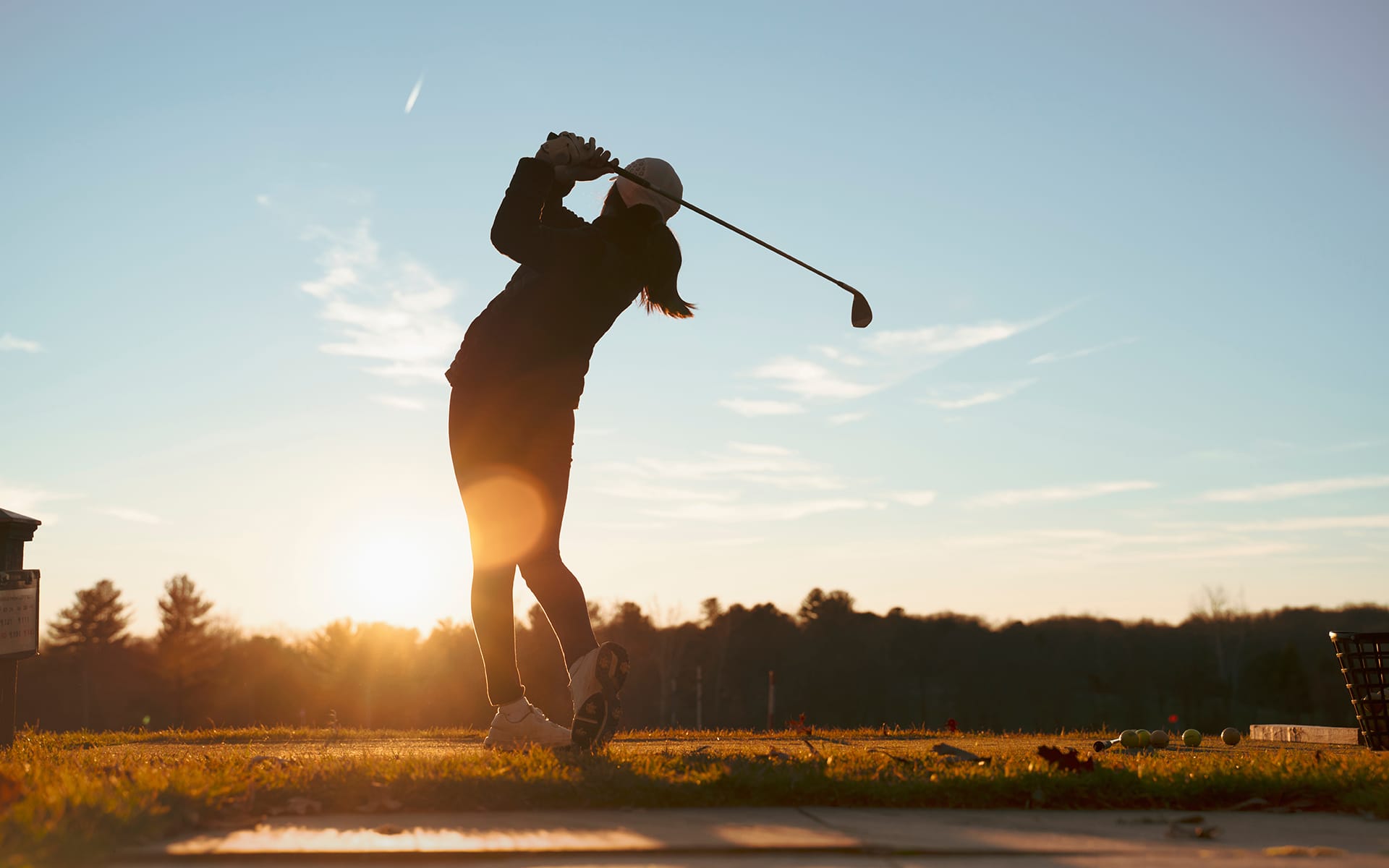 A person swings a golf club on a grassy course at sunset, silhouetted against the sky with trees in the background.