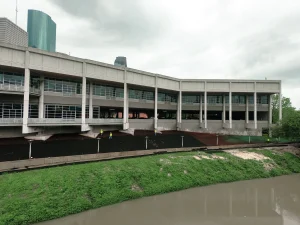 A large concrete building with many windows overlooks a muddy river. Three construction workers in yellow vests are working on dark soil beneath the building. The sky is overcast.