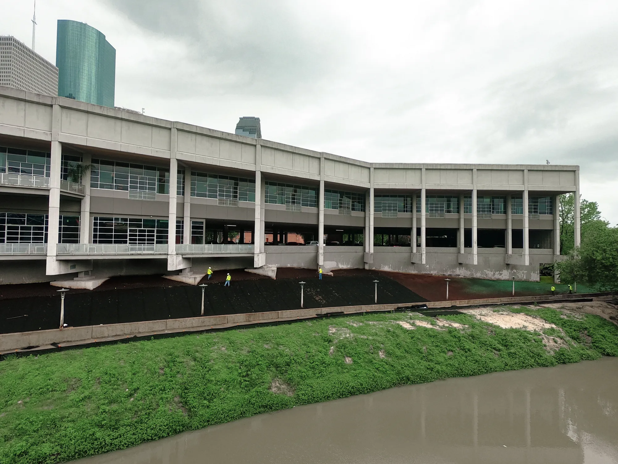 A large concrete building with many windows overlooks a muddy river. Three construction workers in yellow vests are working on dark soil beneath the building. The sky is overcast.