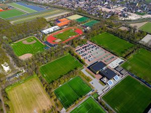 Aerial view of a large sports complex with multiple soccer fields, a baseball diamond, a track and field arena, tennis courts, parking lots, and surrounding green areas near a residential neighborhood.