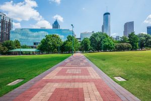 A brick pathway runs through a green park toward city buildings and skyscrapers under a bright blue sky with scattered clouds.